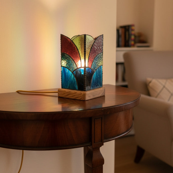 A stained glass table lamp with a geometric, fan-like Art Deco design, resting on a light wood base. The glass panels feature shades of burgundy red, olive green, dark blue, and black, separated by dark metal solder lines.