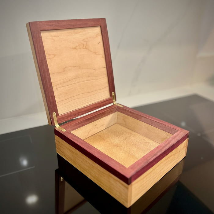 An open wooden keepsake box with a light natural wood interior and reddish-brown outer frame, shown on a dark reflective surface against a light marble-like background.