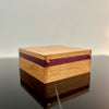 A square wooden keepsake box with a light natural wood grain finish and dark reddish-brown inlay stripes, resting on a dark reflective surface.