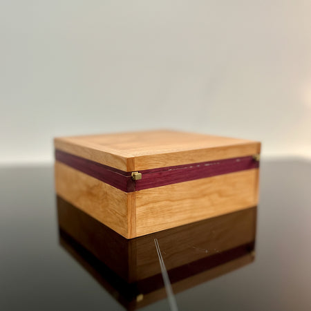 A square wooden keepsake box with a light natural wood grain finish and dark reddish-brown inlay stripes, resting on a dark reflective surface.