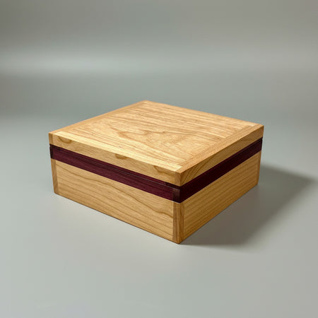 A square wooden box with a light natural wood grain finish and a dark reddish-brown accent stripe along the front edge, shown on a dark reflective surface.
