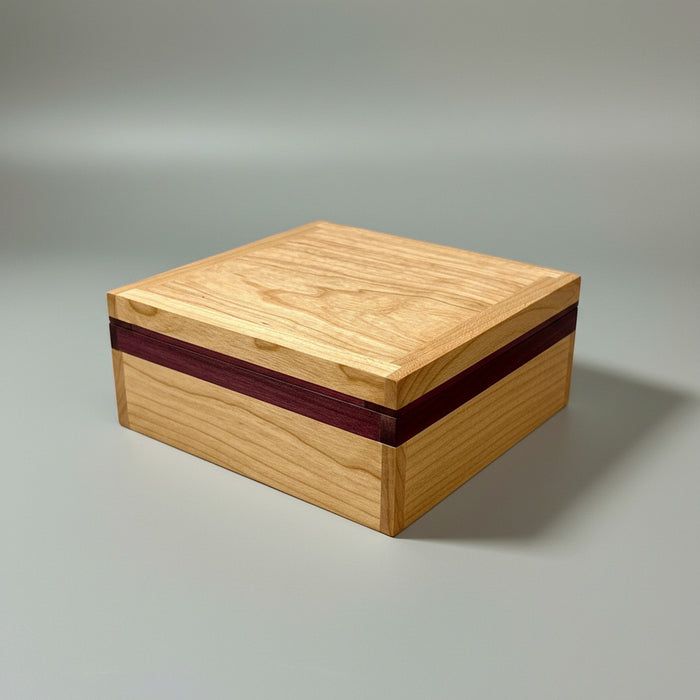 A square wooden box with a light natural wood grain finish and a dark reddish-brown accent stripe along the front edge, shown on a dark reflective surface.