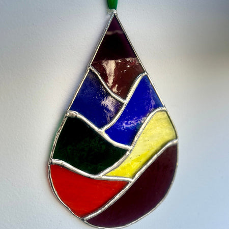 A stained glass ornament in the shape of a teardrop, featuring a colorful abstract pattern with segments of dark purple, blue, green, yellow, red, and brown separated by silver-colored metal lines, hanging from a green ribbon against a plain white wall.