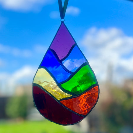 A teardrop-shaped stained glass suncatcher hanging in front of a blue sky with clouds, featuring a rainbow of colored glass panels outlined in black.