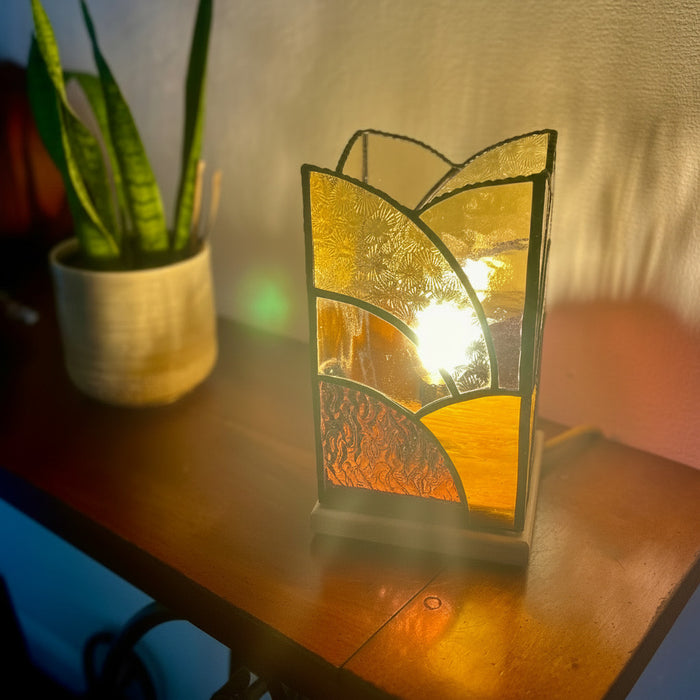 A stained glass pillar table lamp with a warm candle bulb inside, set on a dark wooden table next to a potted snake plant against a textured wall.