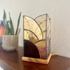 A stained glass table lamp with a geometric art nouveau design in gold, amber, and dark brown tones, resting on a wooden table next to a potted plant.