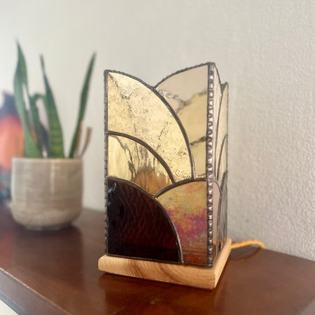 A stained glass table lamp with a geometric art nouveau design in gold, amber, and dark brown tones, resting on a wooden table next to a potted plant.