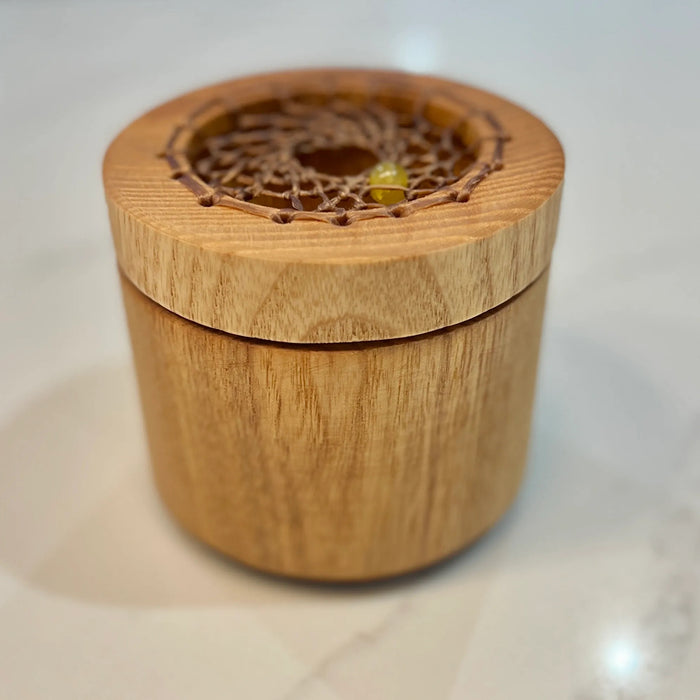 A small cylindrical wooden box with a lid, featuring a circular cutout on top with a delicate woven web design and a tiny yellow bead accent, set on a light marble surface.